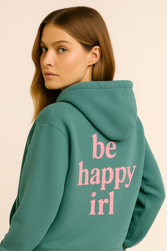 Woman wearing a teal hoodie with 'be happy irl' text on the back, standing against a beige background.